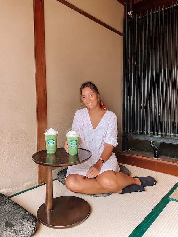 A woman sitting on a tatami floor holding a matcha cup, and another cup in a small round table