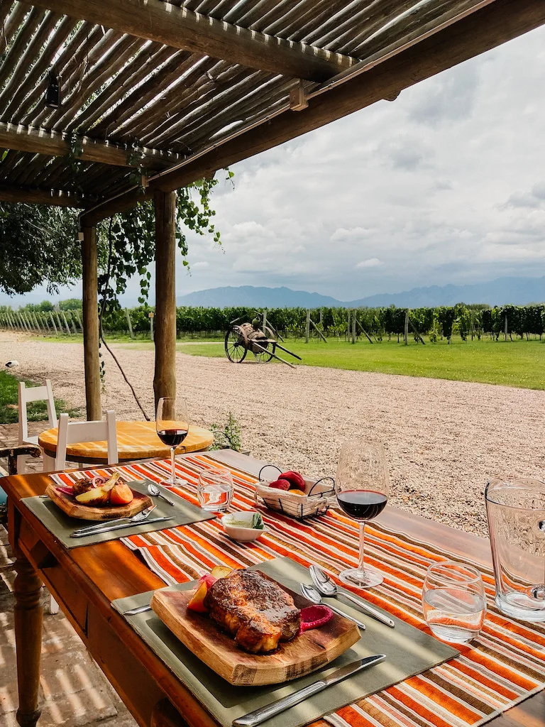 A rustic outdoor dining setup at a winery in Mendoza, Argentina. A wooden table with striped placemats holds grilled steak, fresh bread, and glasses of red wine, with a scenic vineyard and mountains in the background