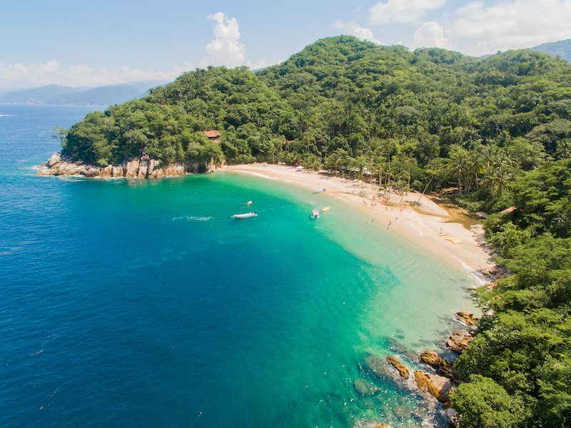 A drone image of a beach with turquoise waters, golden sand, and lush vegetation behind it