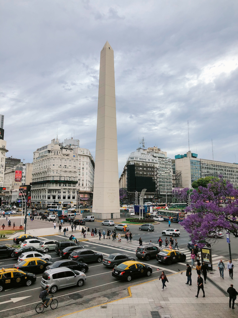 The Buenos Aires Obelisk surrounded by busy traffic, people crossing, and buildings 