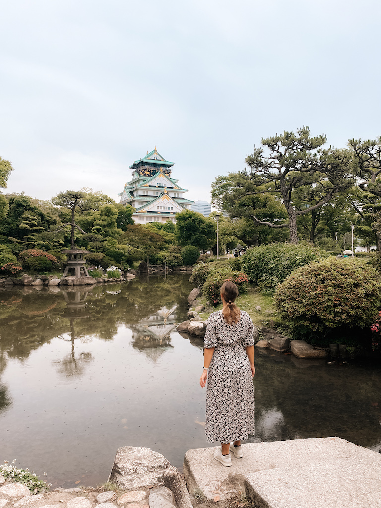 A woman standing on the shores of a lake, and a Japanese building on the other side 