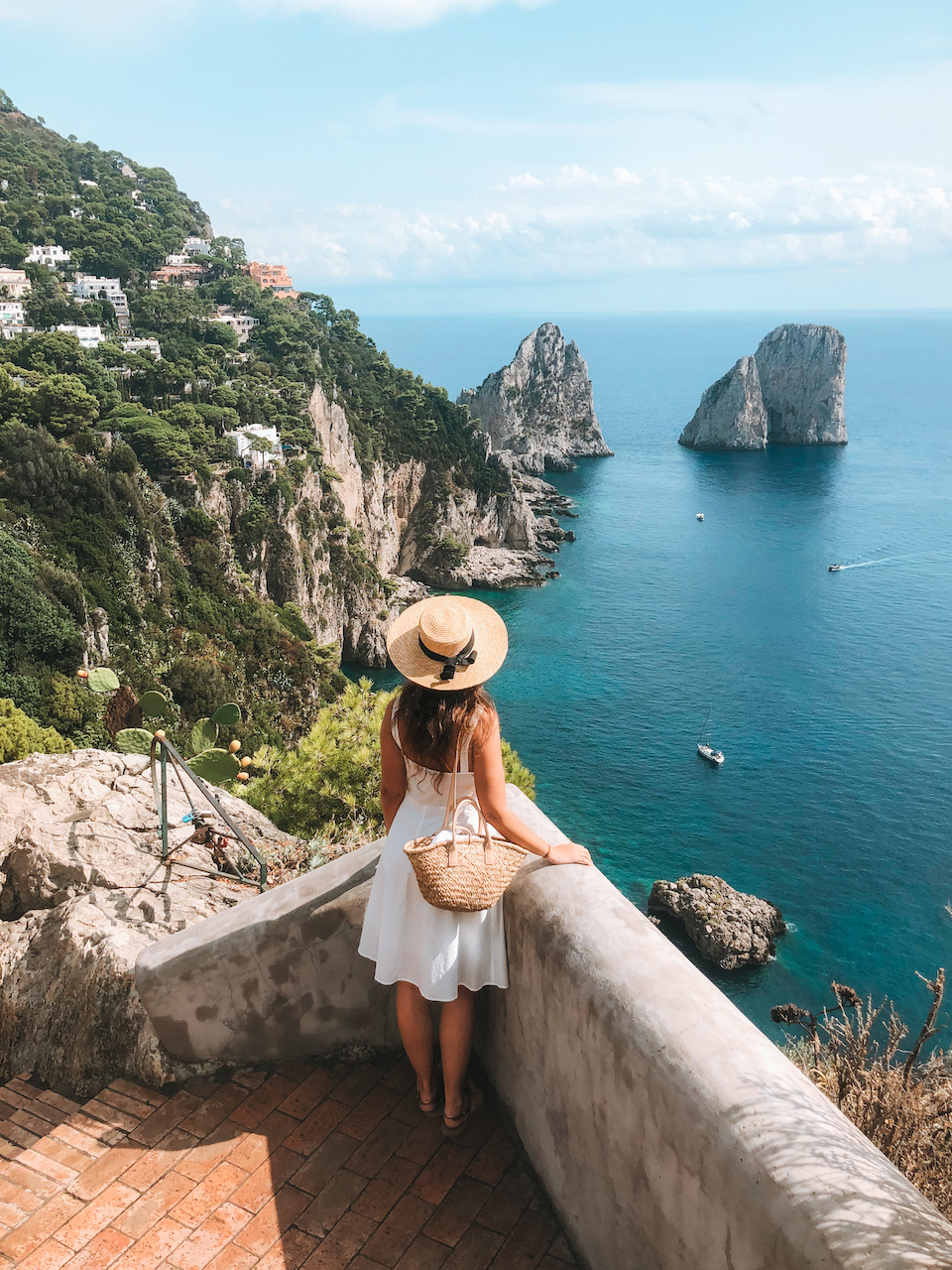 A woman wearing a white dress, a straw hat, and a purse standing in a viewpoint overlooking the Faraglioni rock formations and the sea