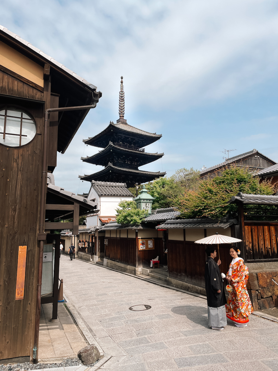 A Japanese couple dressed in traditional attire, with the man holding a white paper umbrella, are standing in a traditional alley in Kyoto. In the background stands the Yasaka Pagoda.