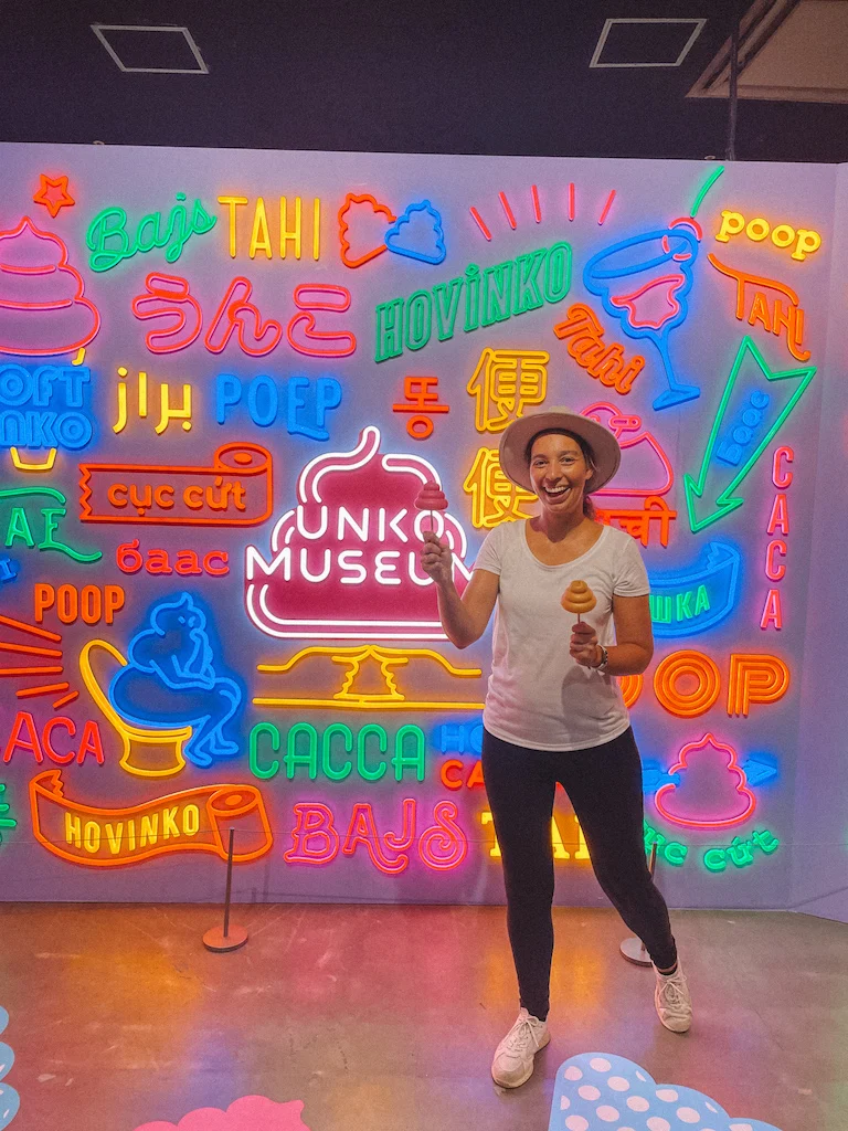 A woman holding poop-shaped lollipops in front of a wall with neon signs