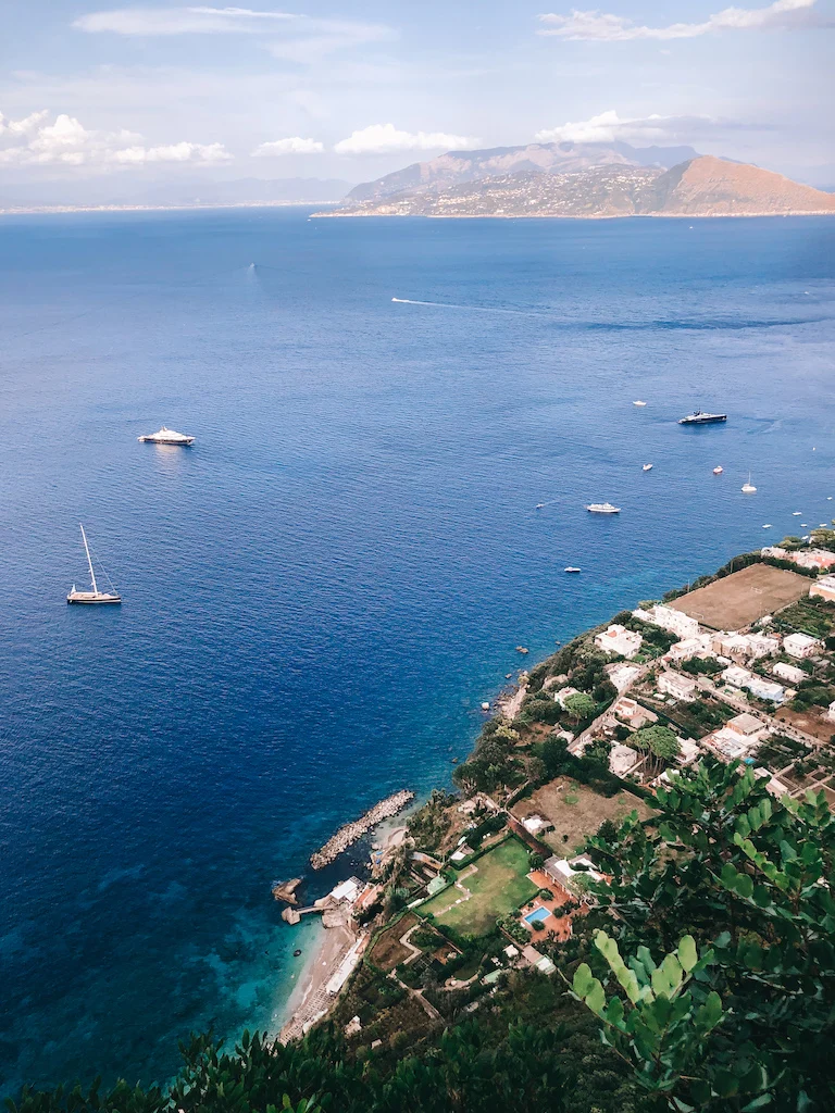 An aerial image of the sea with a few bots in it, and land on the right with vegetation and a few houses