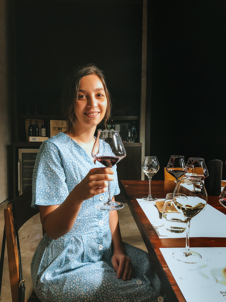 A woman in a light blue dress holding a glass of red wine and smiling at the camera in a winery during a wine tour in Mendoza, Argentina.