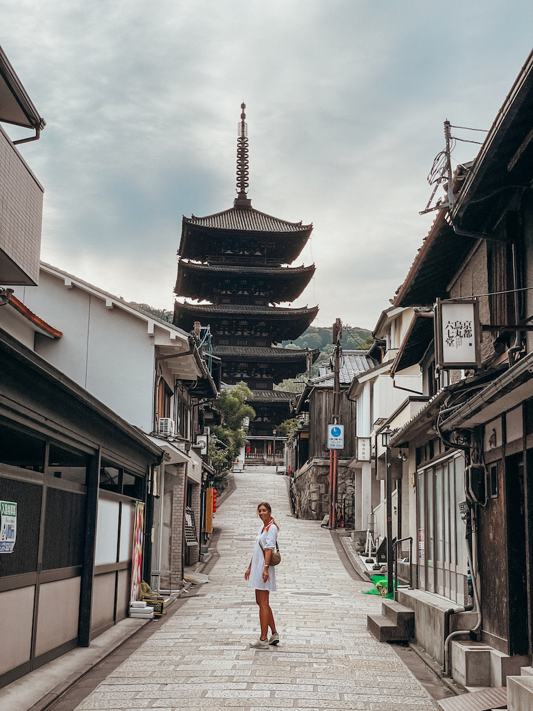 A woman with a white dress is smiling at the camera and standing in front of the Yasaka Pagoda in Kyoto.