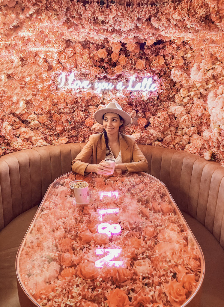 A woman having coffee on a table that's fully decorated with pink flowers, and behind her a wall full of flowers with a neon sign that reads ''I love you a latte''