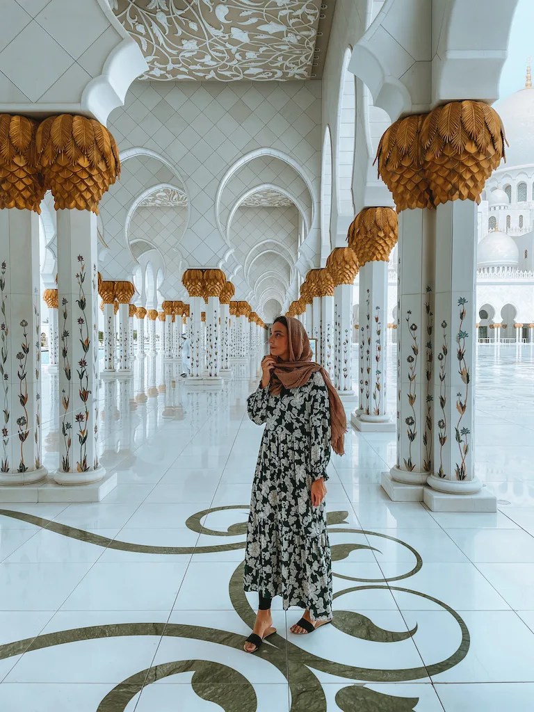 A woman in a long floral dress and headscarf standing in the elegant white and gold corridor of Sheikh Zayed Grand Mosque, surrounded by intricate columns.