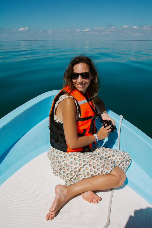 A woman wearing a life jacket and sunglasses sits barefoot at the front of a small blue boat, smiling as she enjoys a peaceful boat tour in the Sian Ka'an Biosphere Reserve.