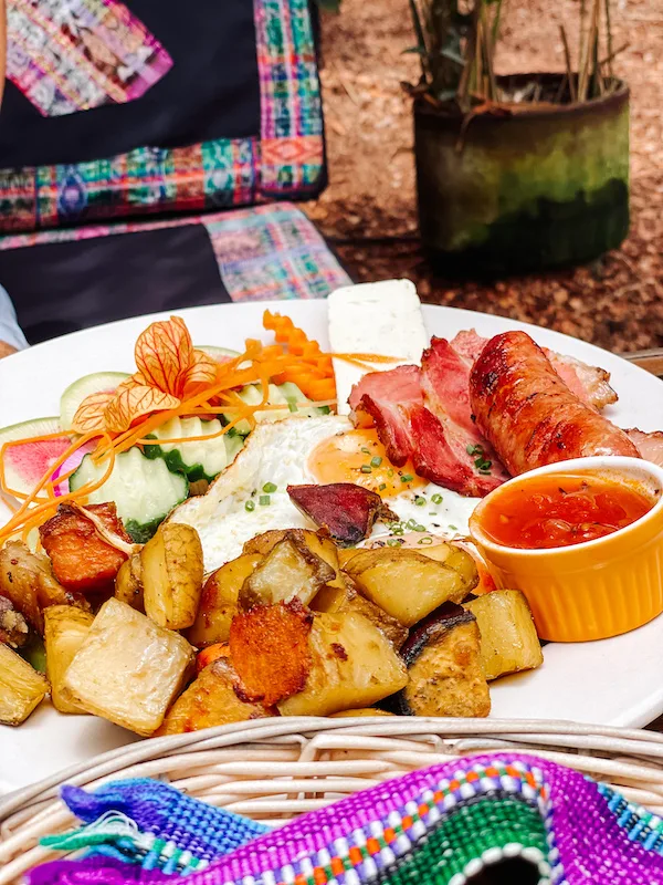 A close-up image of a place full of food, including potatoes, eggs, sausages, bacon, cucumber, and a small bowl with a red sauce