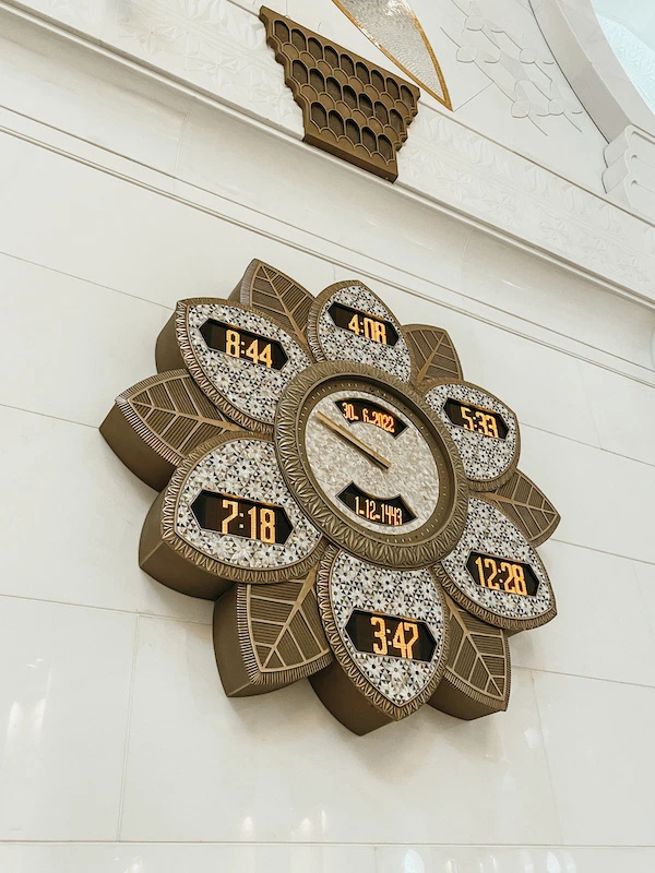 A decorative clock inside Sheikh Zayed Grand Mosque displaying prayer times in Arabic numerals, designed in a floral pattern.