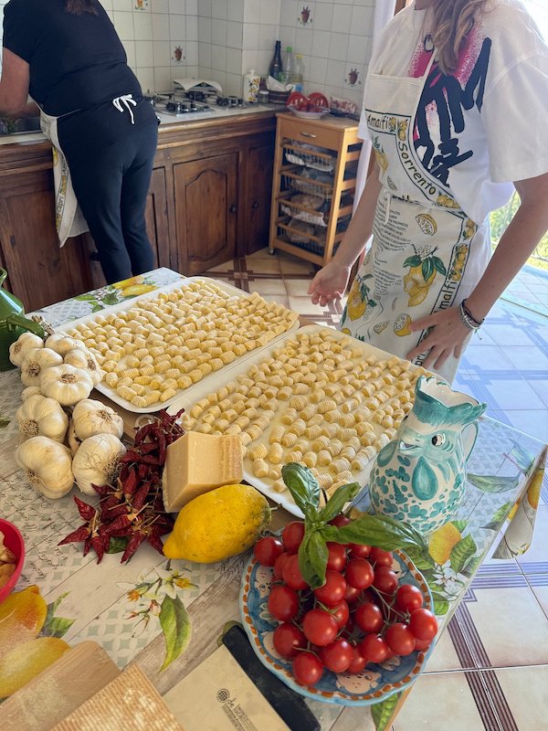 A kitchen counter with trays of fresh gnocchi, tomatoes, garlic, chili peppers, and Parmesan, with a person in an apron preparing food.