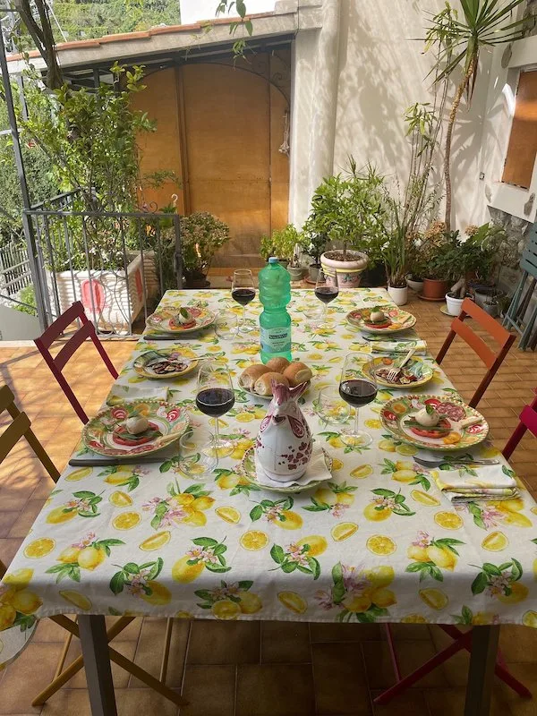 An outdoor dining table in Positano with a lemon-patterned tablecloth, colorful plates, wine glasses, and a decorative pitcher, surrounded by greenery.