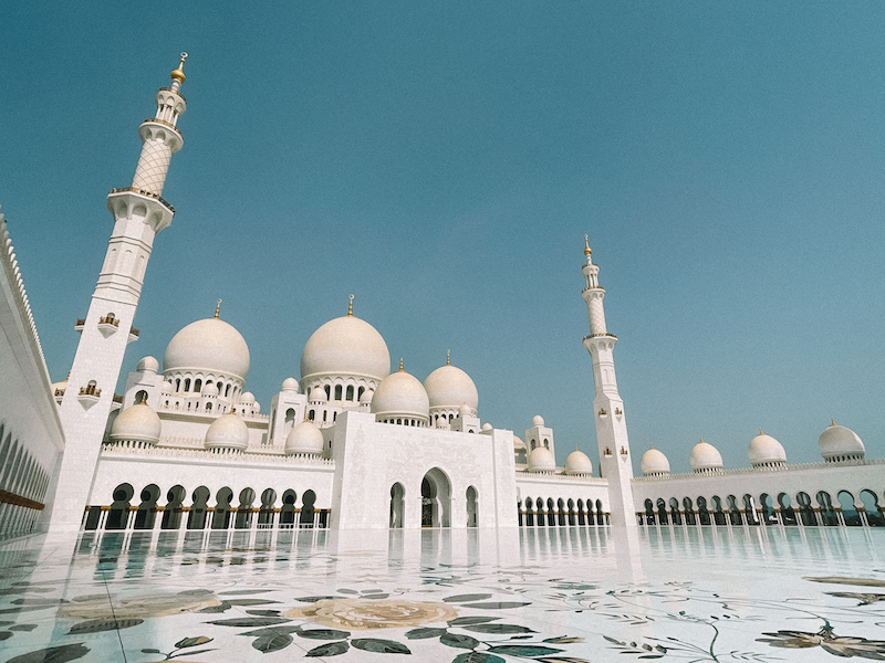 The stunning white marble courtyard of Sheikh Zayed Grand Mosque, reflecting intricate floral designs and surrounded by domes under a bright blue sky.