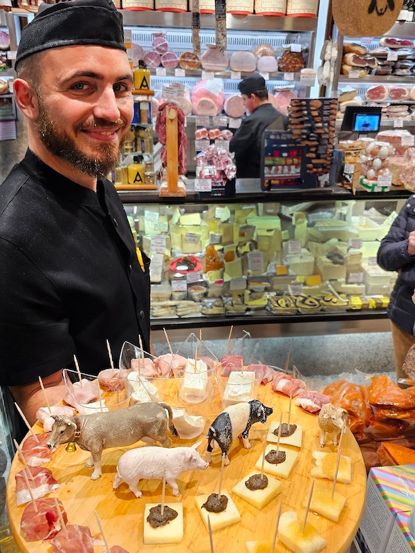 A friendly vendor at a Roman deli presenting a wooden platter filled with cheese and cured meats, each topped with a miniature farm animal figurine to indicate its origin. Behind him, a well-stocked counter displays an array of cheeses and charcuterie.