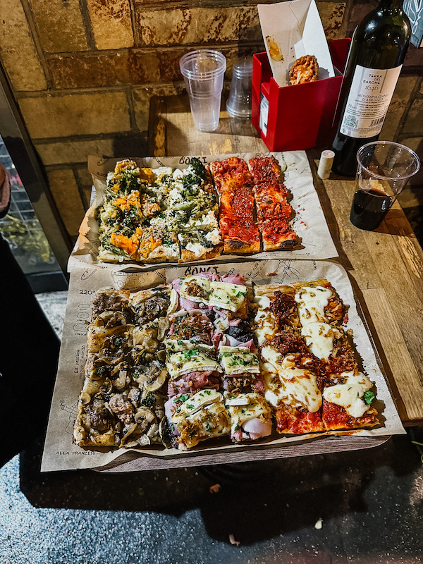 A selection of Roman-style pizza al taglio with various toppings, served on wooden trays at a casual eatery in Rome. A bottle of red wine and a box of fried supplì sit on the table.