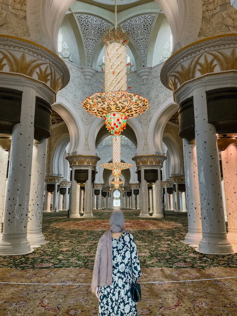 A woman admiring the massive crystal chandelier inside the prayer hall of Sheikh Zayed Grand Mosque, with grand pillars and detailed ceilings.