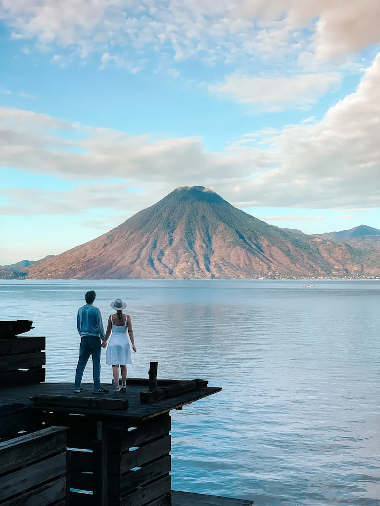 A couple standing on a wooden deck on the shores of a lake, looking at a volcano and holding hands.