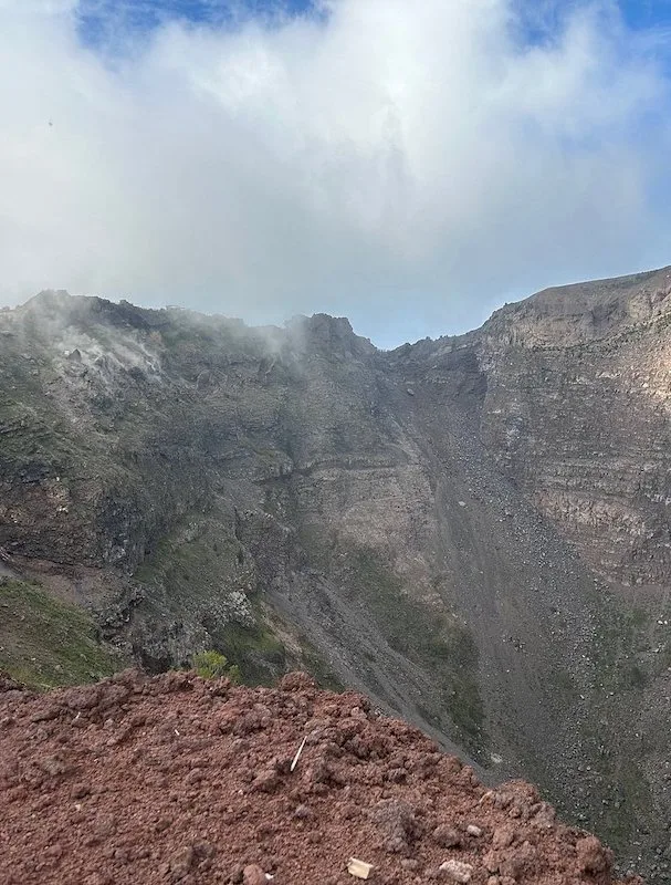 A view of the crater of Mount Vesuvius, with steam rising from the rocky terrain. The dramatic landscape, partially covered in mist, highlights the volcano's active nature.