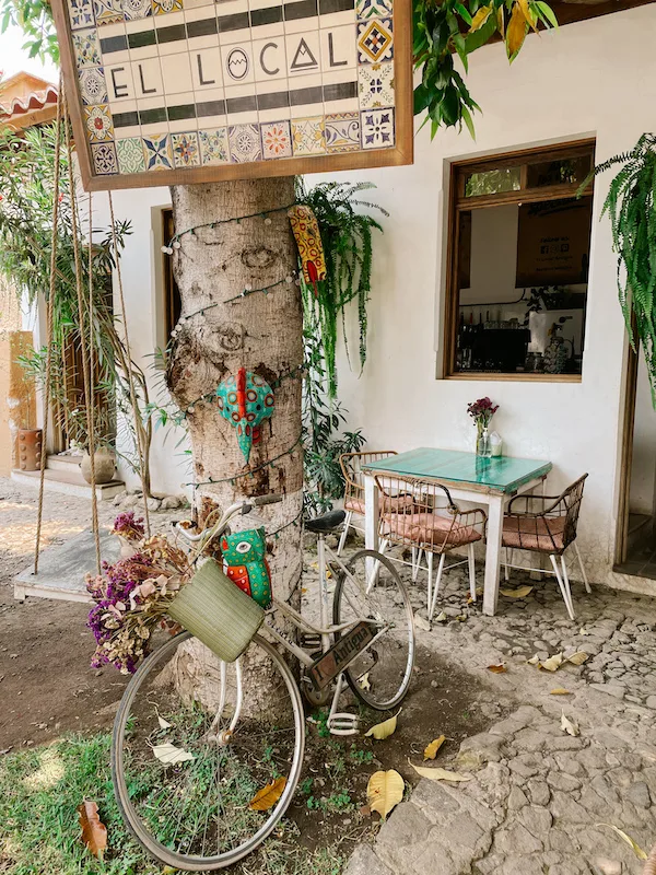 A tree with decorations, a sign that reads ''El Local'' and a bicycle resting on it. There's a small table with chairs in the background 