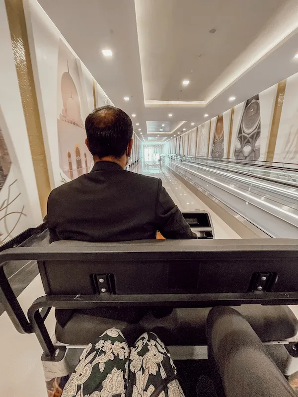 A view from inside a buggy transporting visitors through the entrance corridor of Sheikh Zayed Grand Mosque, lined with elegant white walls and gold details.