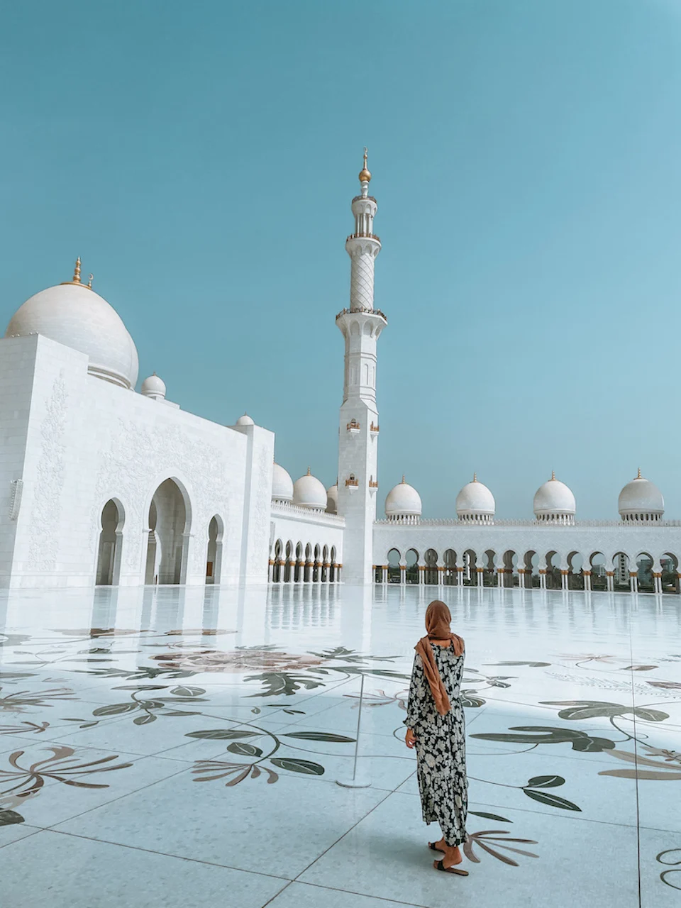 A woman standing in the marble courtyard of Sheikh Zayed Grand Mosque, gazing at the grand domes and minaret reflecting on the polished floor.