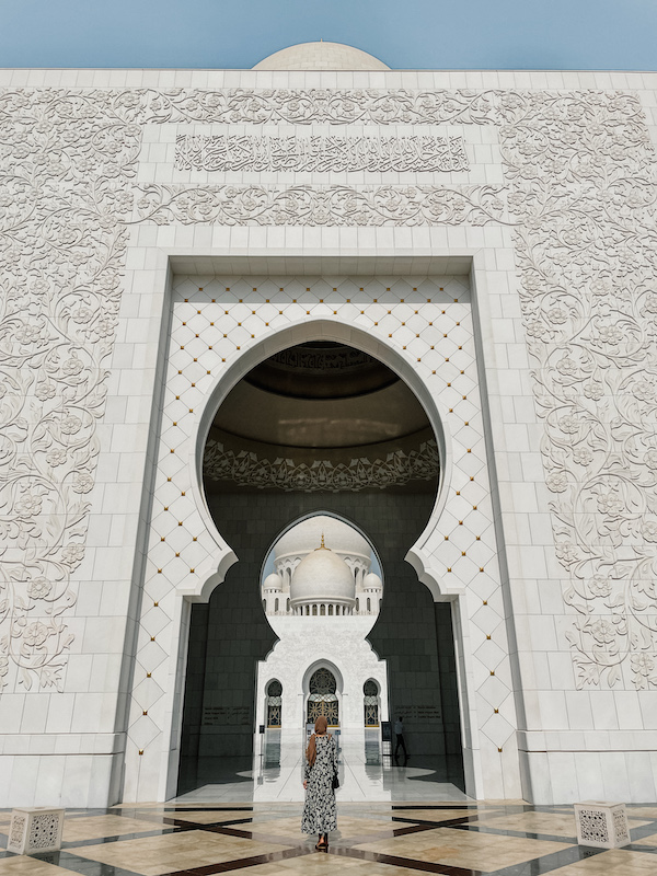 A woman walking toward the grand entrance of Sheikh Zayed Grand Mosque, framed by an ornate white archway with intricate carvings.