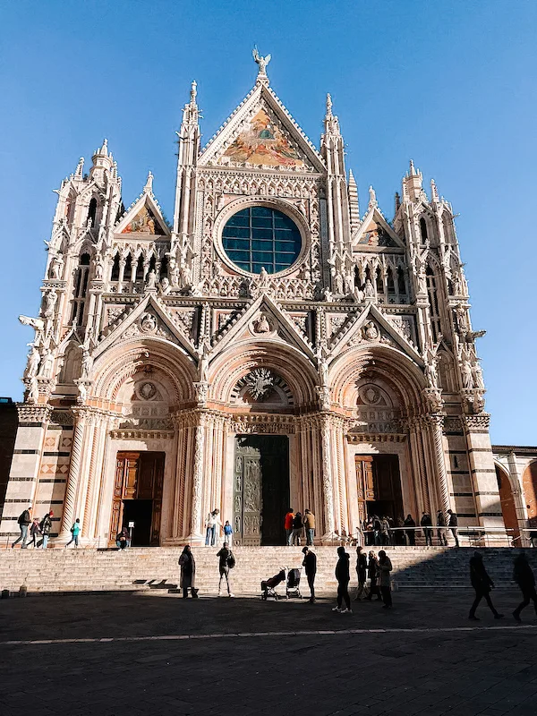 The stunning facade of Siena Cathedral, a masterpiece of Gothic architecture, illuminated by the sun against a clear blue sky. 