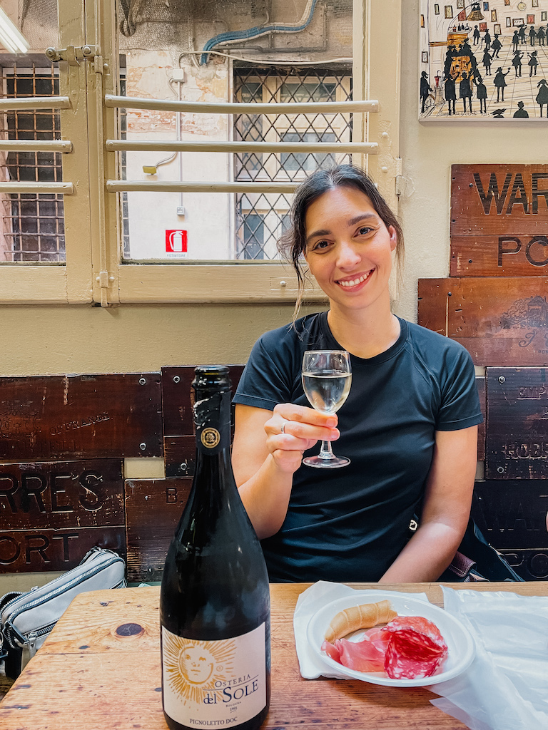 A woman enjoying a glass of white wine at Osteria del Sole in Bologna, with a bottle of Pignoletto and a plate of salami and bread.