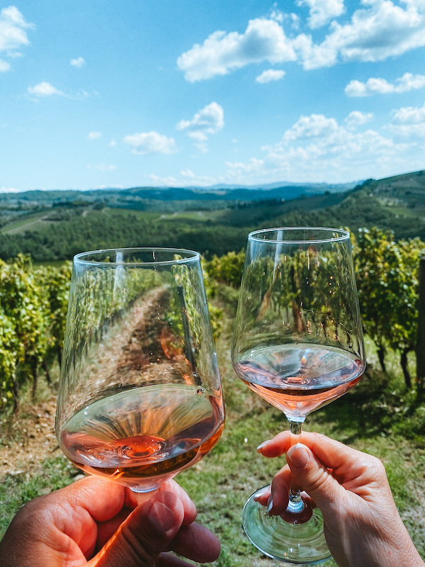 Two hands holding glasses of rosé wine against the backdrop of rolling vineyards in the Tuscan countryside during a wine tour from Florence.